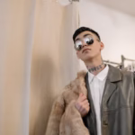 Stylish man posing indoors with fur coat and sunglasses, showcasing modern fashion.