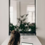 A woman with curly hair works on a laptop, facing a window with plants, symbolizing remote work.
