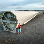A massive wind turbine blade lying outdoors with people nearby for scale.